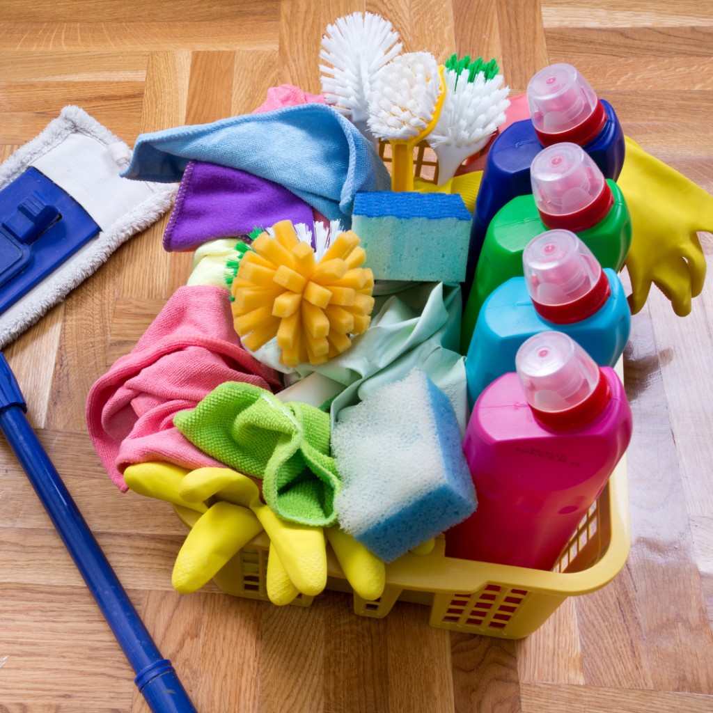 Basket of colorful detergents, cloths, and cleaning tools beside a mop